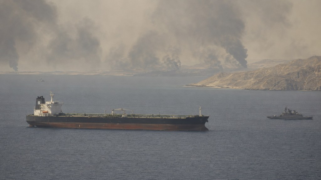 Petrolero navegando en aguas del estrecho de Ormuz con un buque militar y columnas de humo sobre la costa.